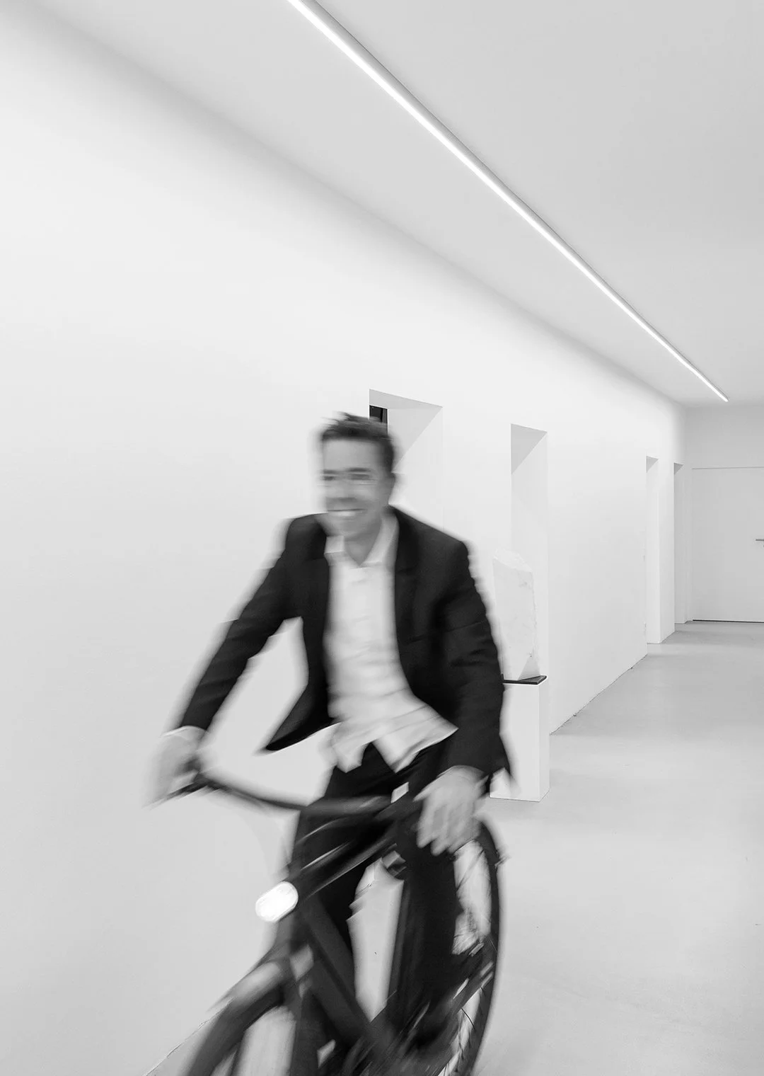 Ein lachender Mann in einem schwarzen Anzug fährt Fahrrad in einem weißen, minimalistischen Raum.