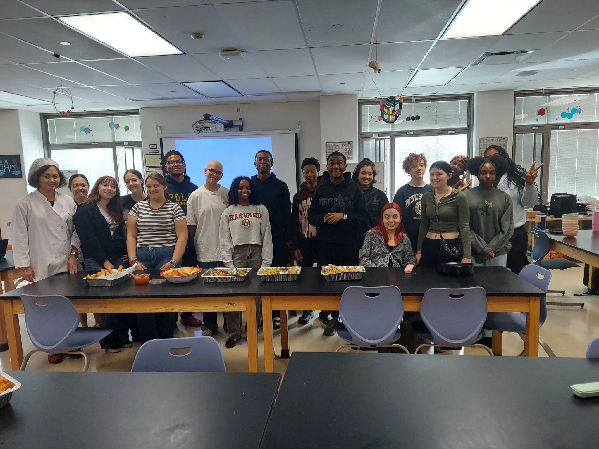 A diverse group of young adults and a teacher of Blesame culinary class in a classroom standing behind tables with trays of food, smiling at the camera.