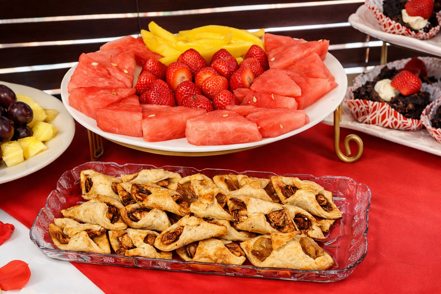 Fruit platter with sliced watermelon, strawberries, and mango slices, with a tray of baklava in front and chocolate desserts with strawberries on a separate platter to the right.