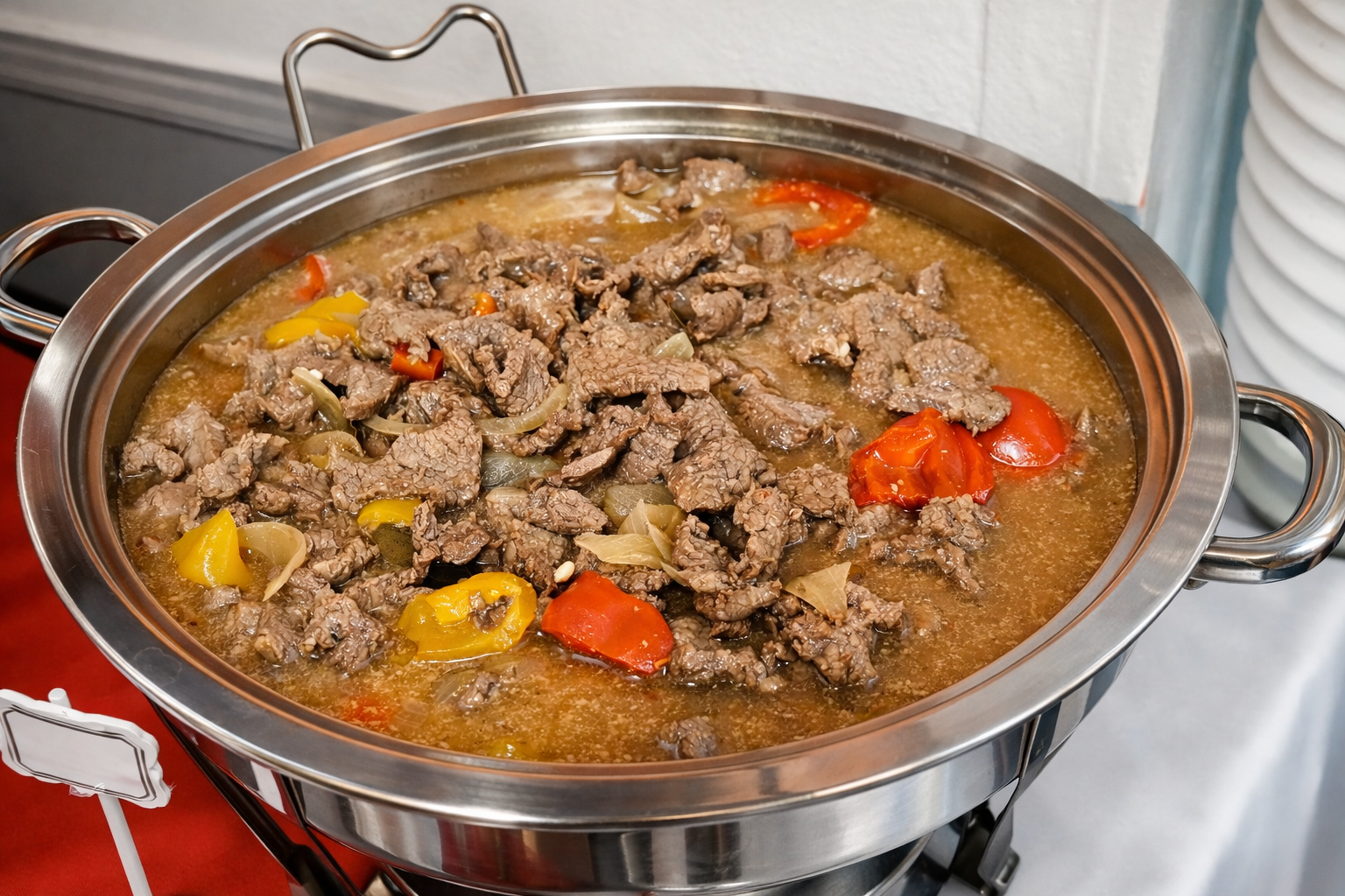 A large stainless steel chafing dish containing beef stew with chunks of beef, red and yellow bell peppers, onions, and a brown broth.