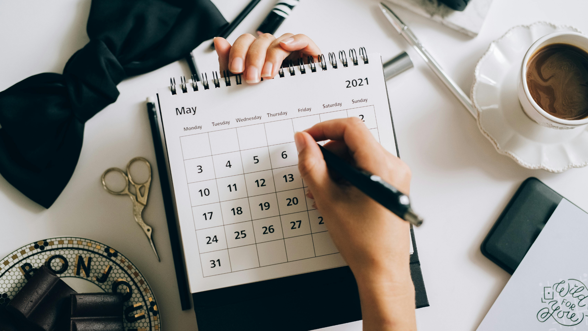 A person writing on a May 2021 desk calendar with a black marker. The desk has a pair of small scissors, a computer mouse, and a coffee cup on a decorative saucer.