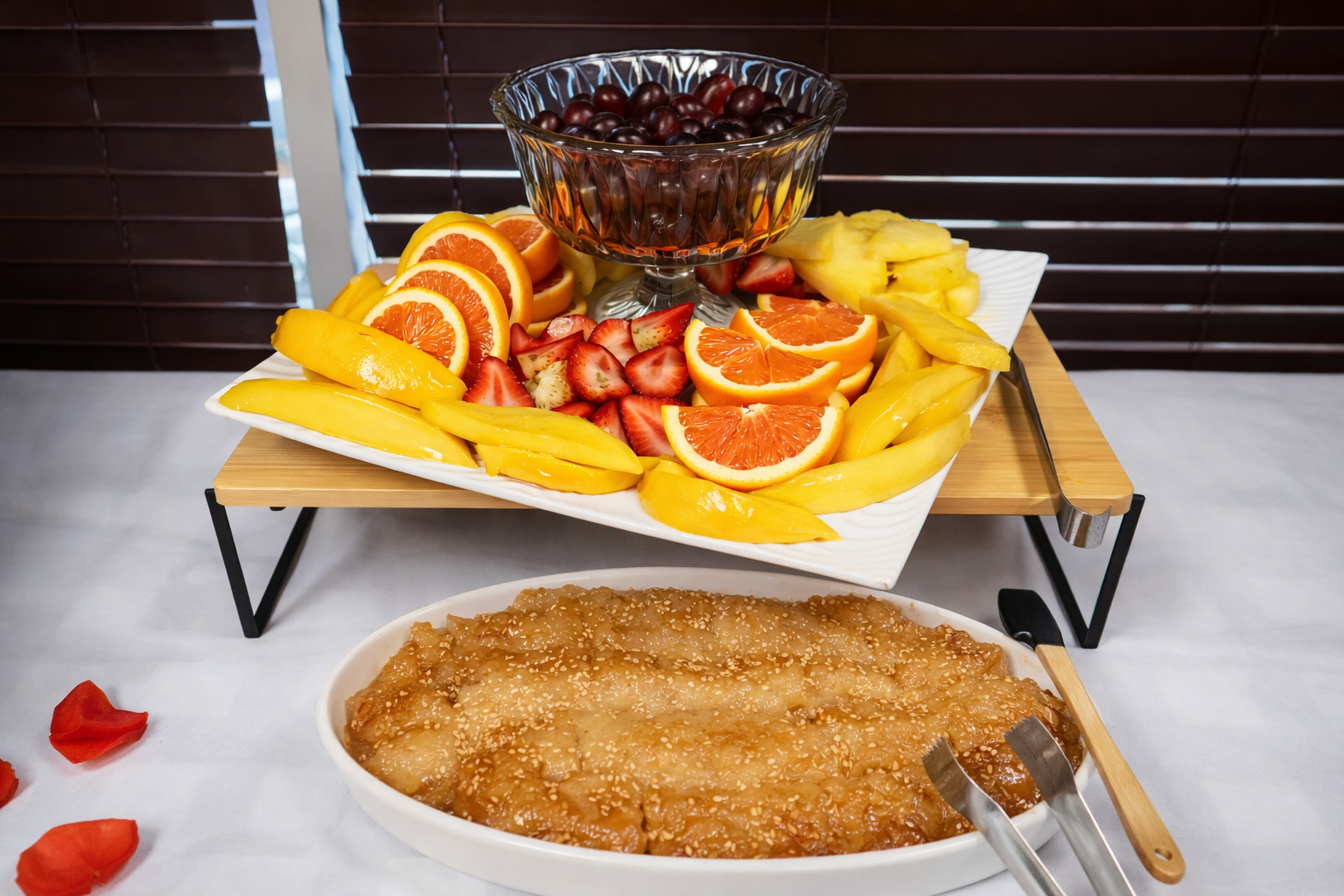 A fruit display on a two-tiered stand with sliced oranges, strawberries, mangoes, and a bowl of grapes, accompanied by a large dish of cooked apples with sesame seeds.