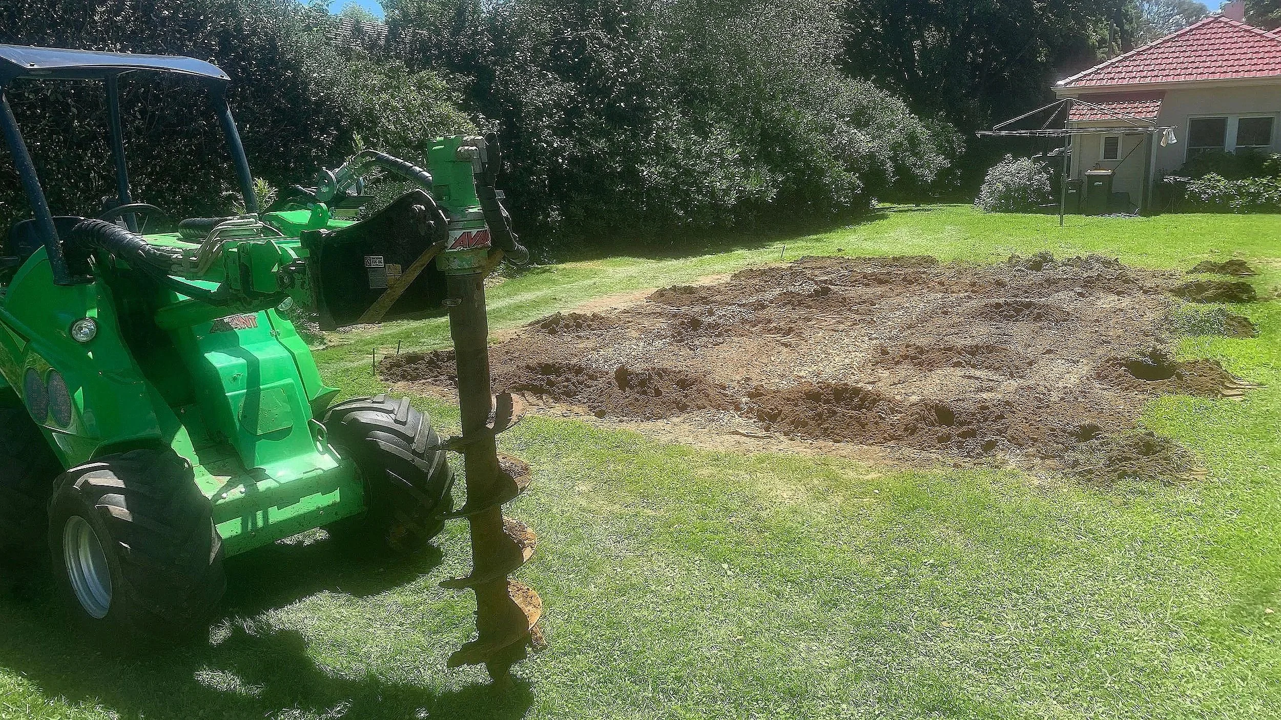 A small green tractor with an auger attachment is digging a hole in a grassy backyard, with a house and trees in the background.