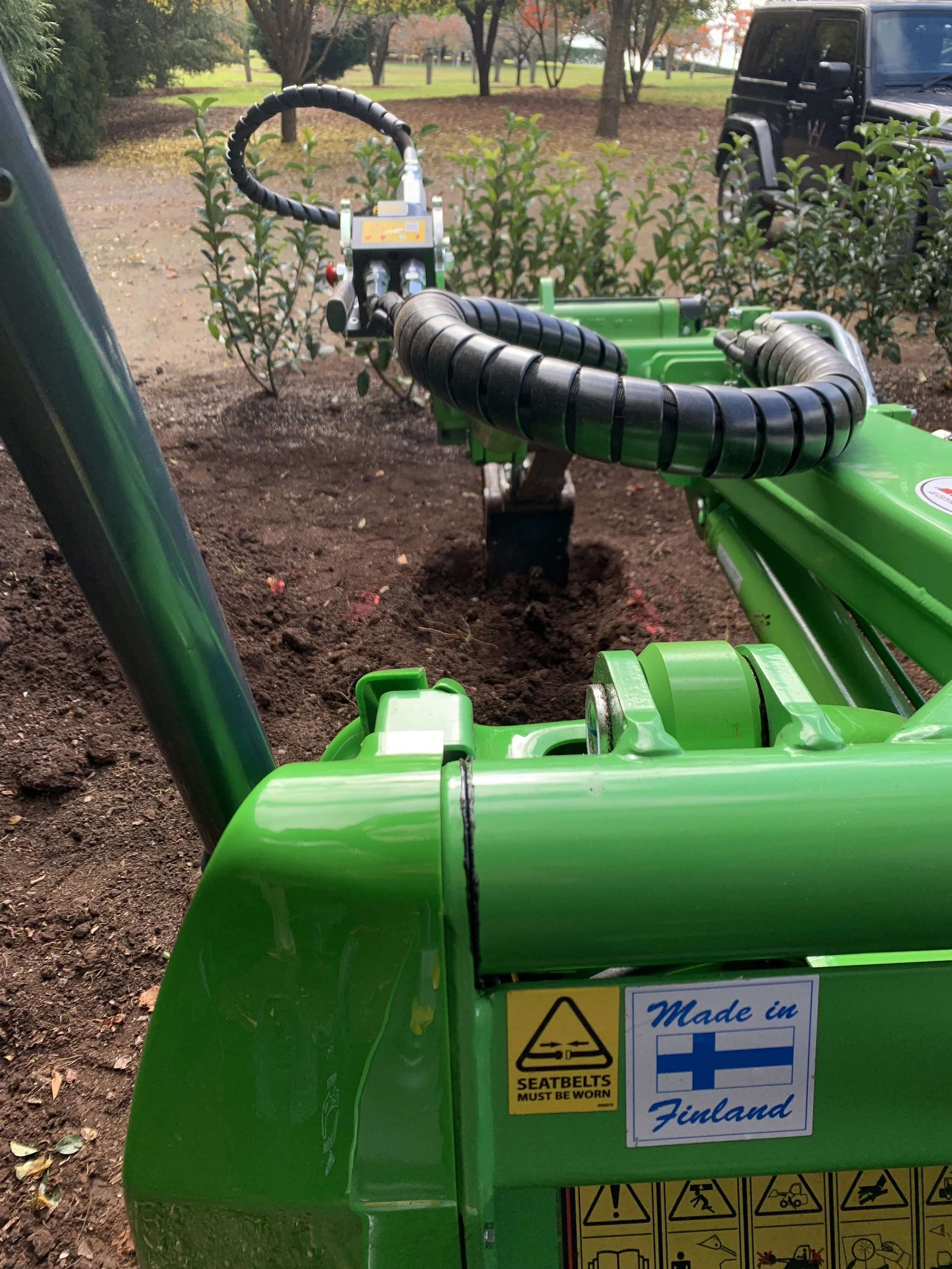 green Avant loader with digger attachement digging a hole in soil with green bush in the background