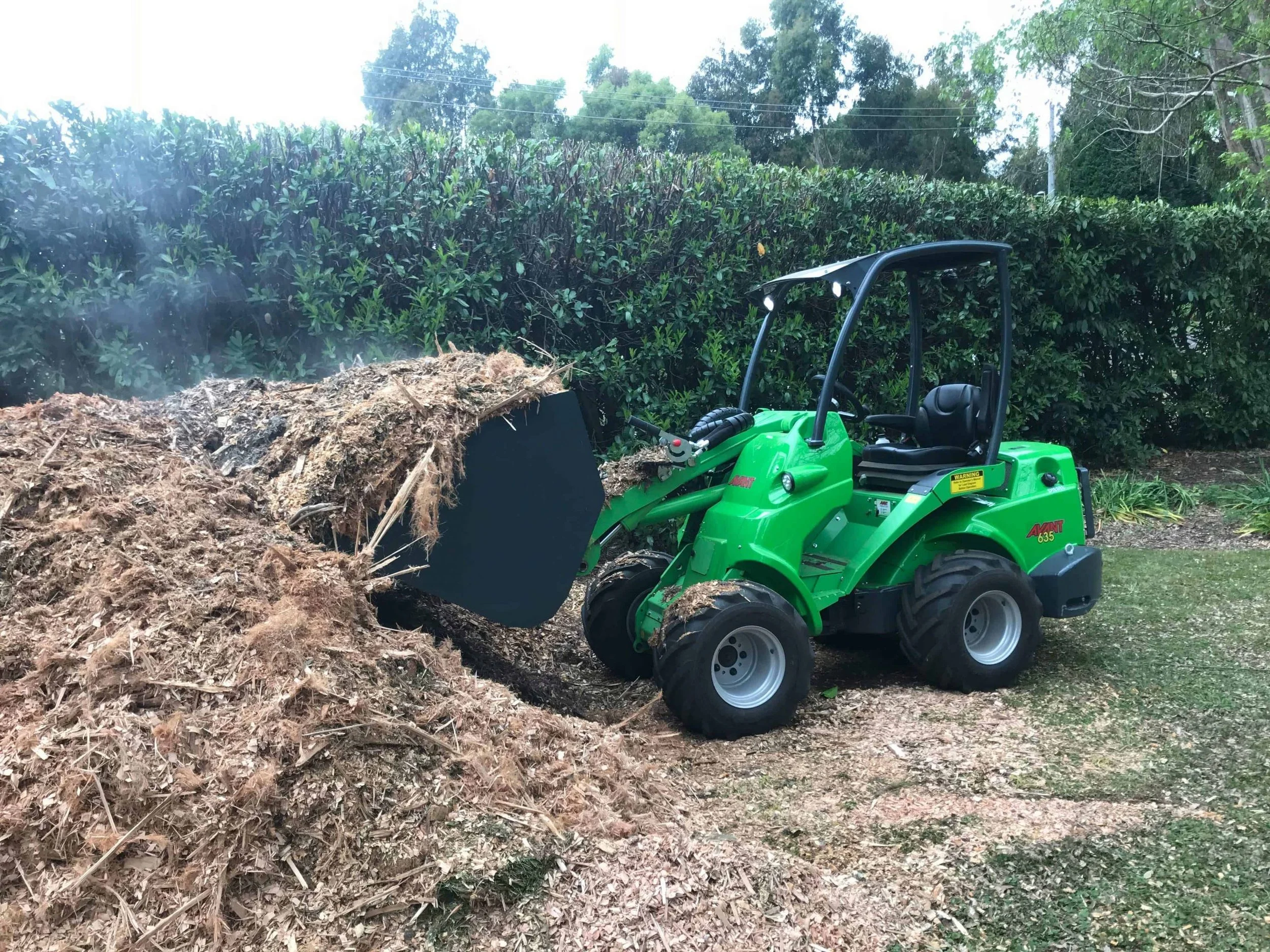 green Avant loader with general bucket loading mulch with green bush in the background