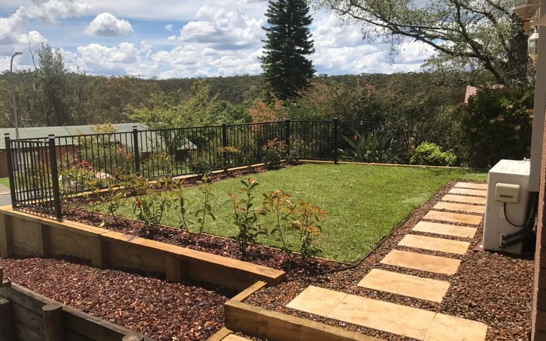 A backyard with a small lawn, a pathway of stepping stones, and a black security fence. There are trees and hills in the background under a partly cloudy sky.