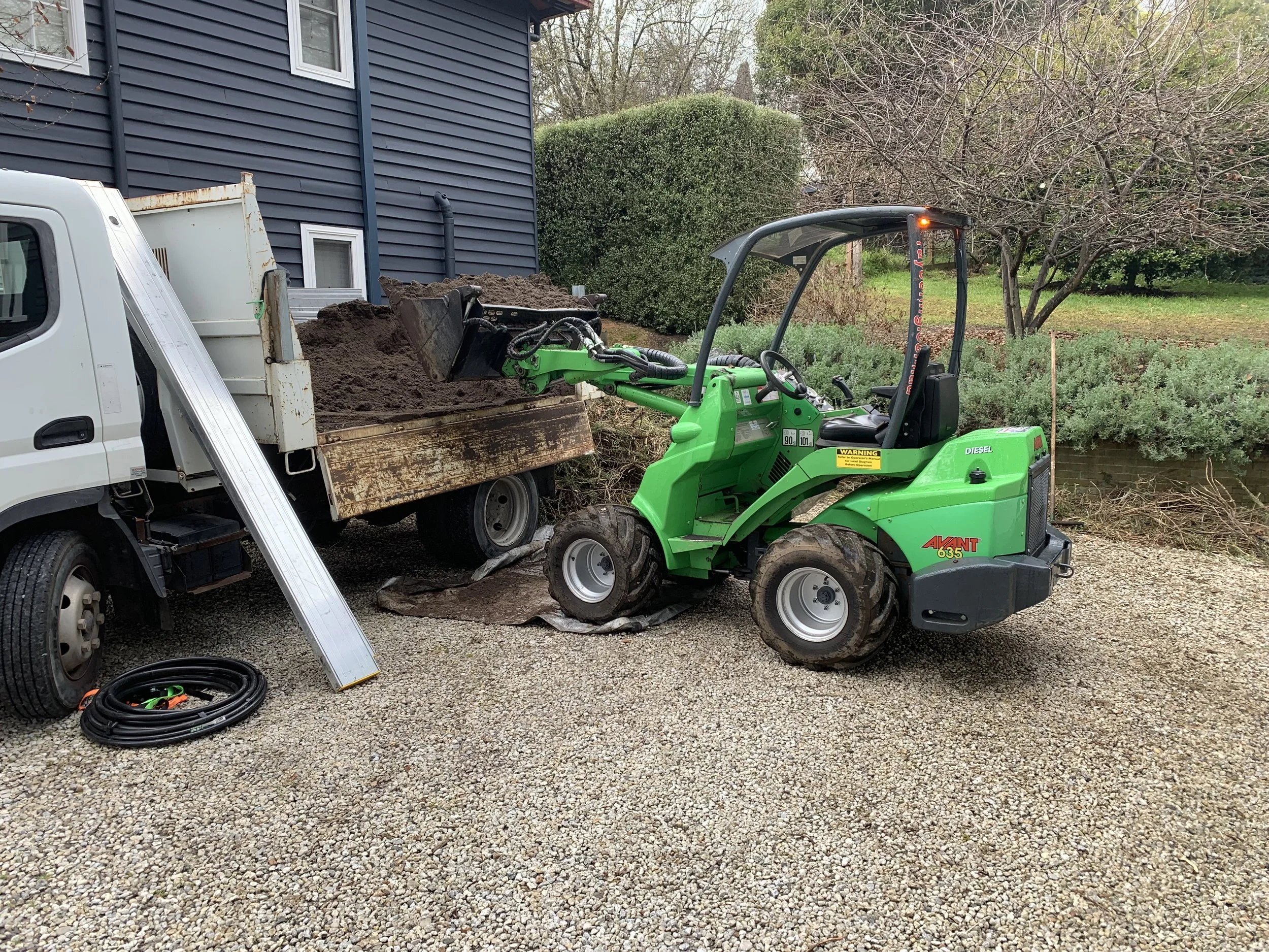 A green mini front loader scooping dirt from a truck bed beside a house with blue siding, surrounded by trees and bushes.