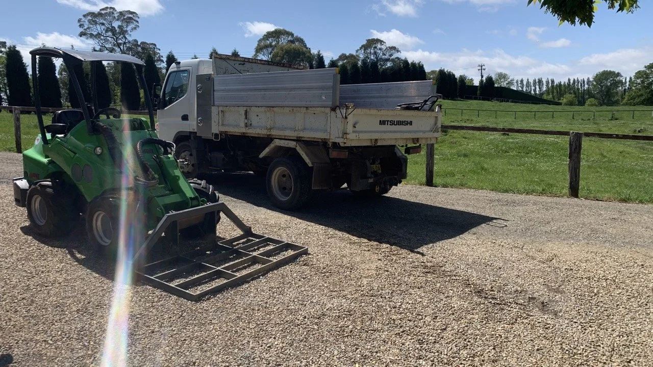 green loader with level bar attachment on driveway with pebbles  with a truck behind and paddock in the background
