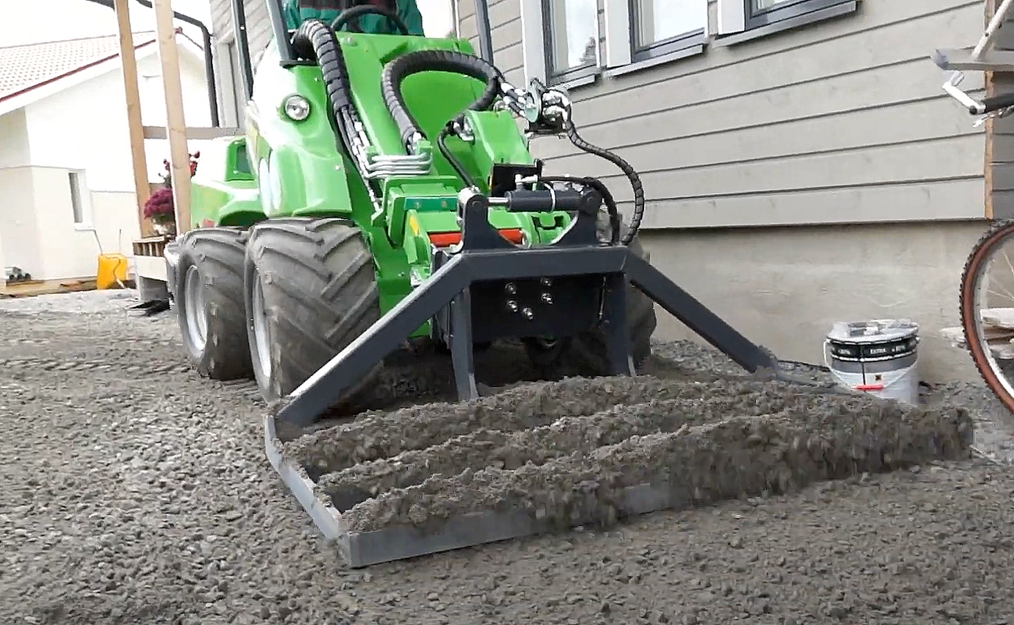 A green compact loader with large black tires working on a gravelly surface outdoors next to a house with beige siding.