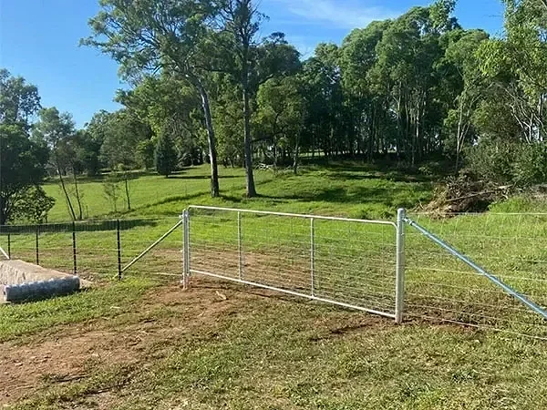 Stock fence with a metal gate opening to a rural farmland area, surrounded by trees and clear sky in the distance