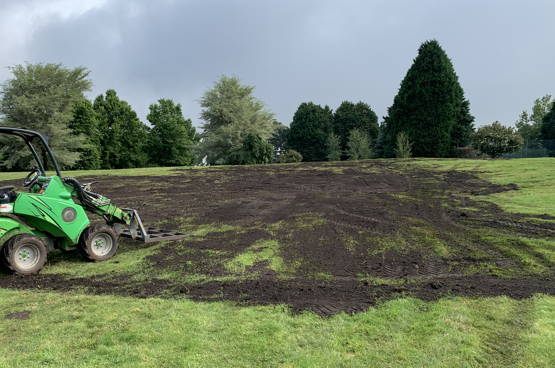 A green tractor with a front loader attachment working on a patch of soil in a grassy field, with trees and a cloudy sky in the background.