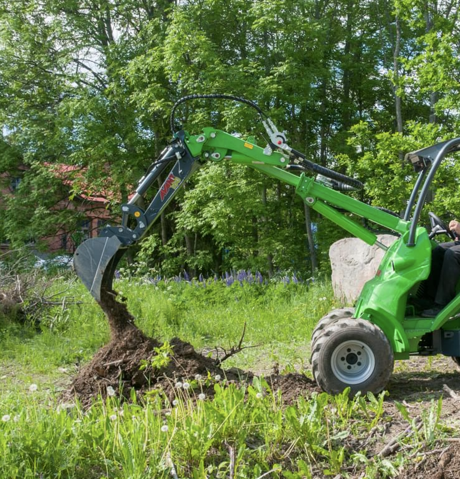 green Avant loader with digger attachment digging soil out with green trees in the background