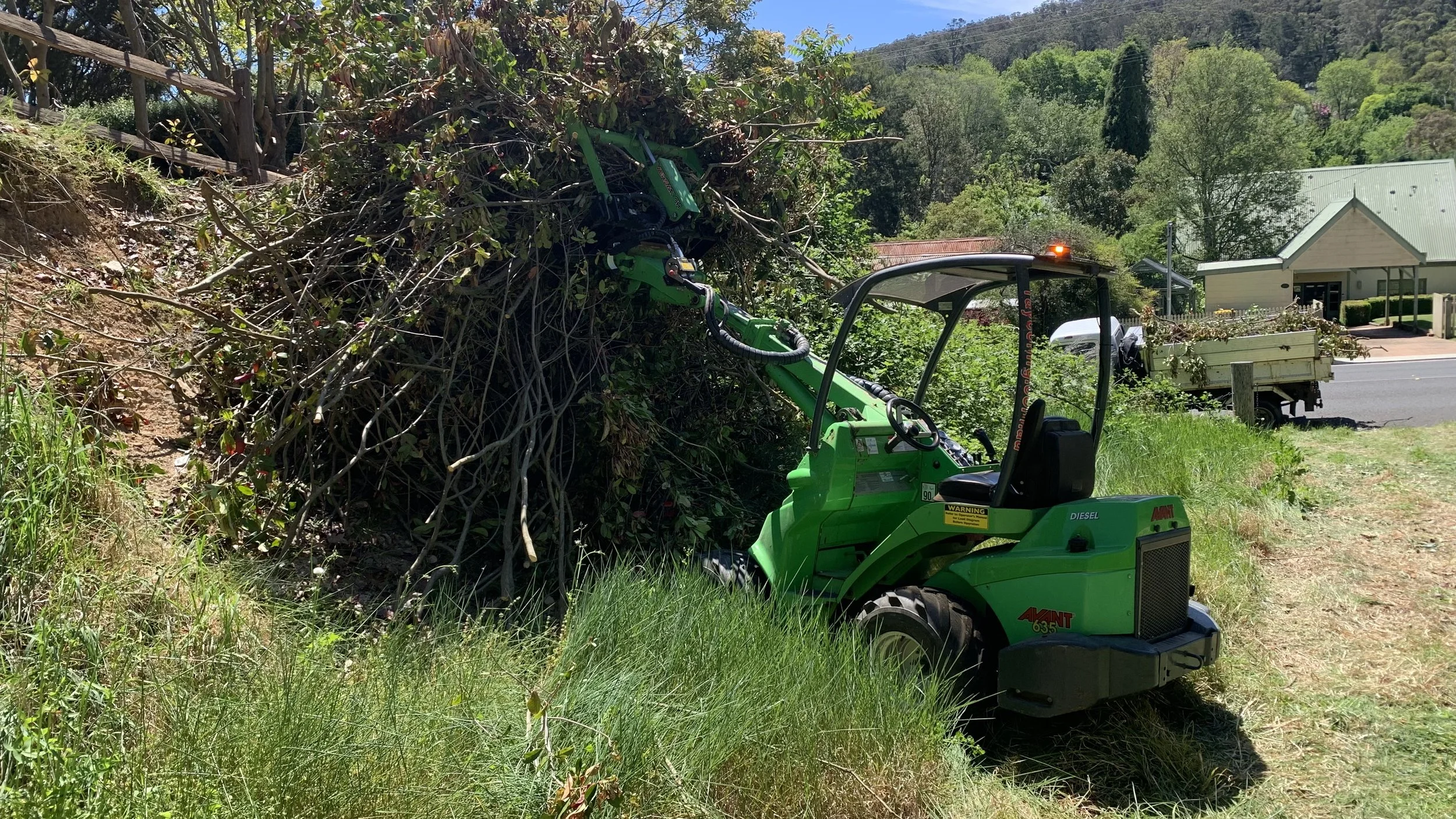 Green compact loader moving branches and brush in an outdoor area with trees, grass, and houses in the background.