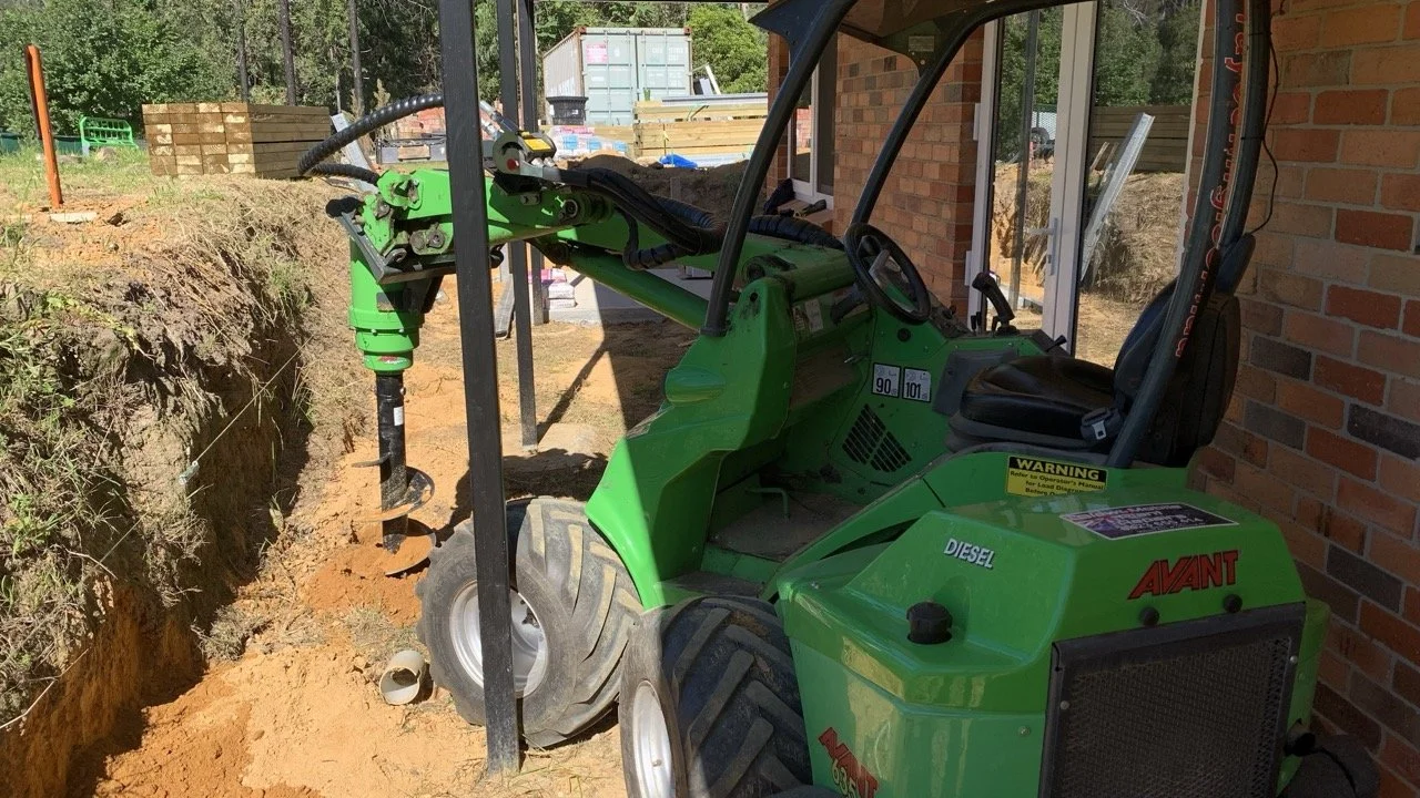 green Avant loader with auger attachment drilling holes with timber fence in the background