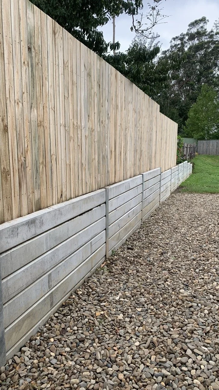 retaining wall with concrete sleepers and timber paling fence on top of it. pebbles on the ground