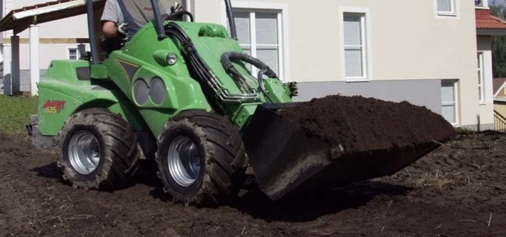 green Avant loader with 4in1 bucket full of soil driving on dirt with white house in the background