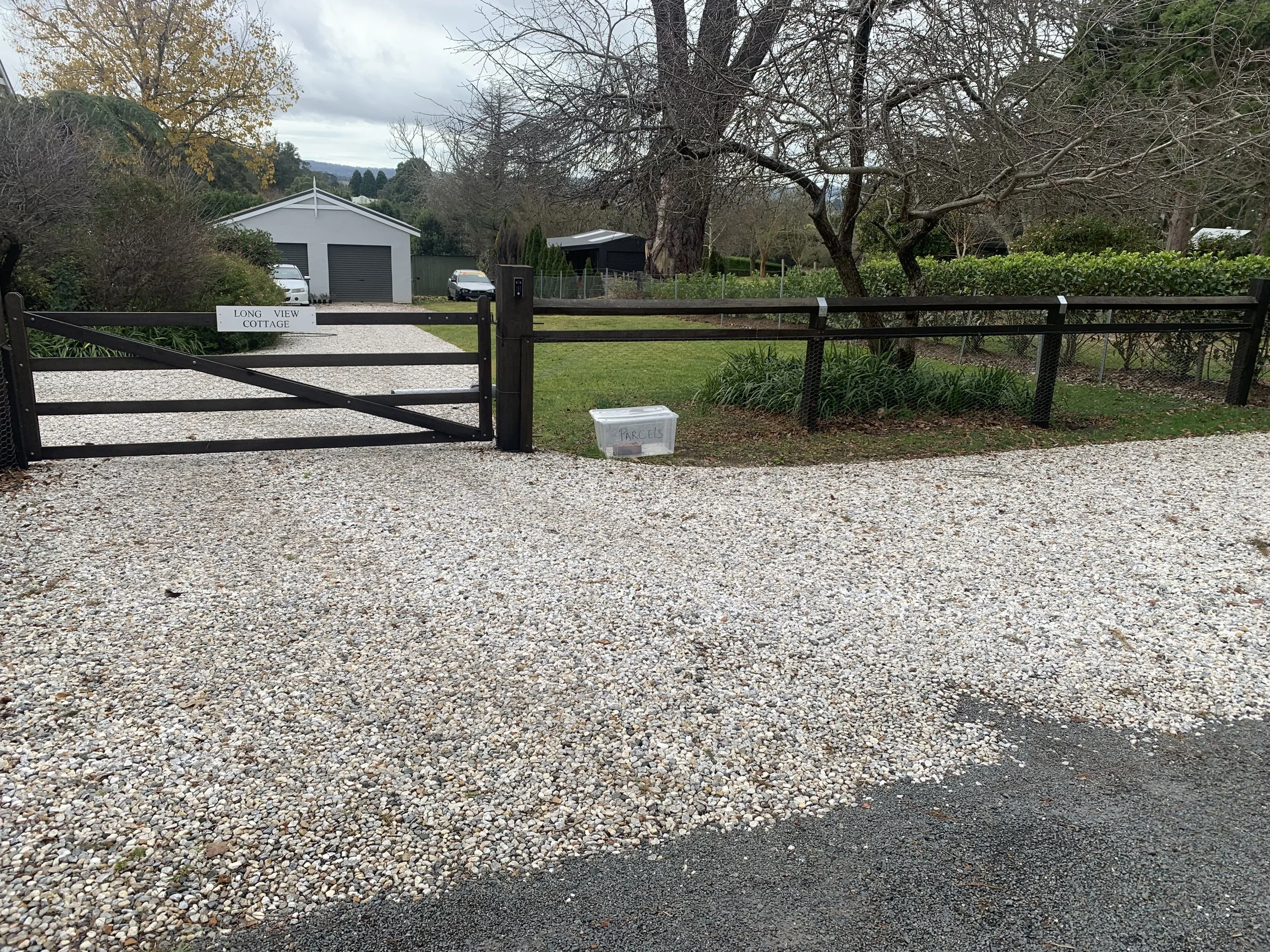 Gravel driveway with wooden property gate, surrounded by well-maintained lawn and trees, leading to a residential property.
