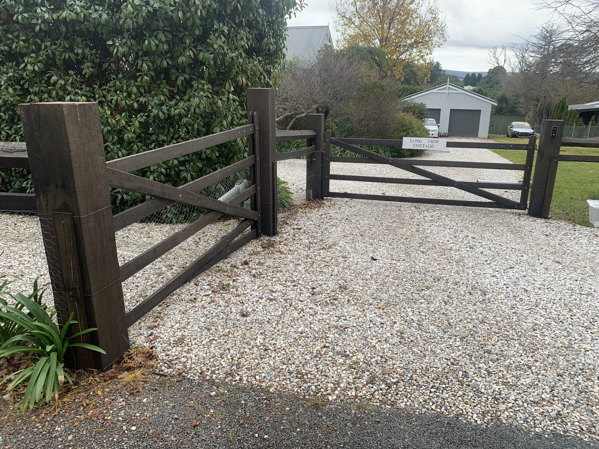 Wooden entrance gate with gravel driveway and signage, leading to a property with well-maintained greenery on both sides.