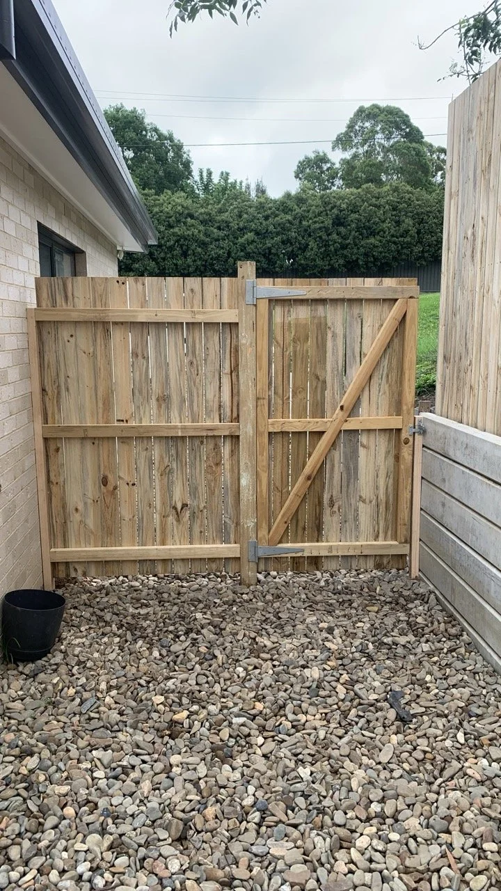 the back of a paling fence with open gate and retaining all with concrete sleepers and pebbles on the ground