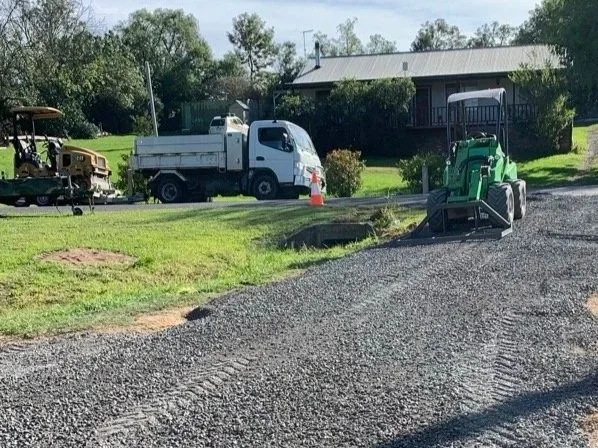 Green machin Avant loader, spreading road base with level bar attachment with truck and roller in the background