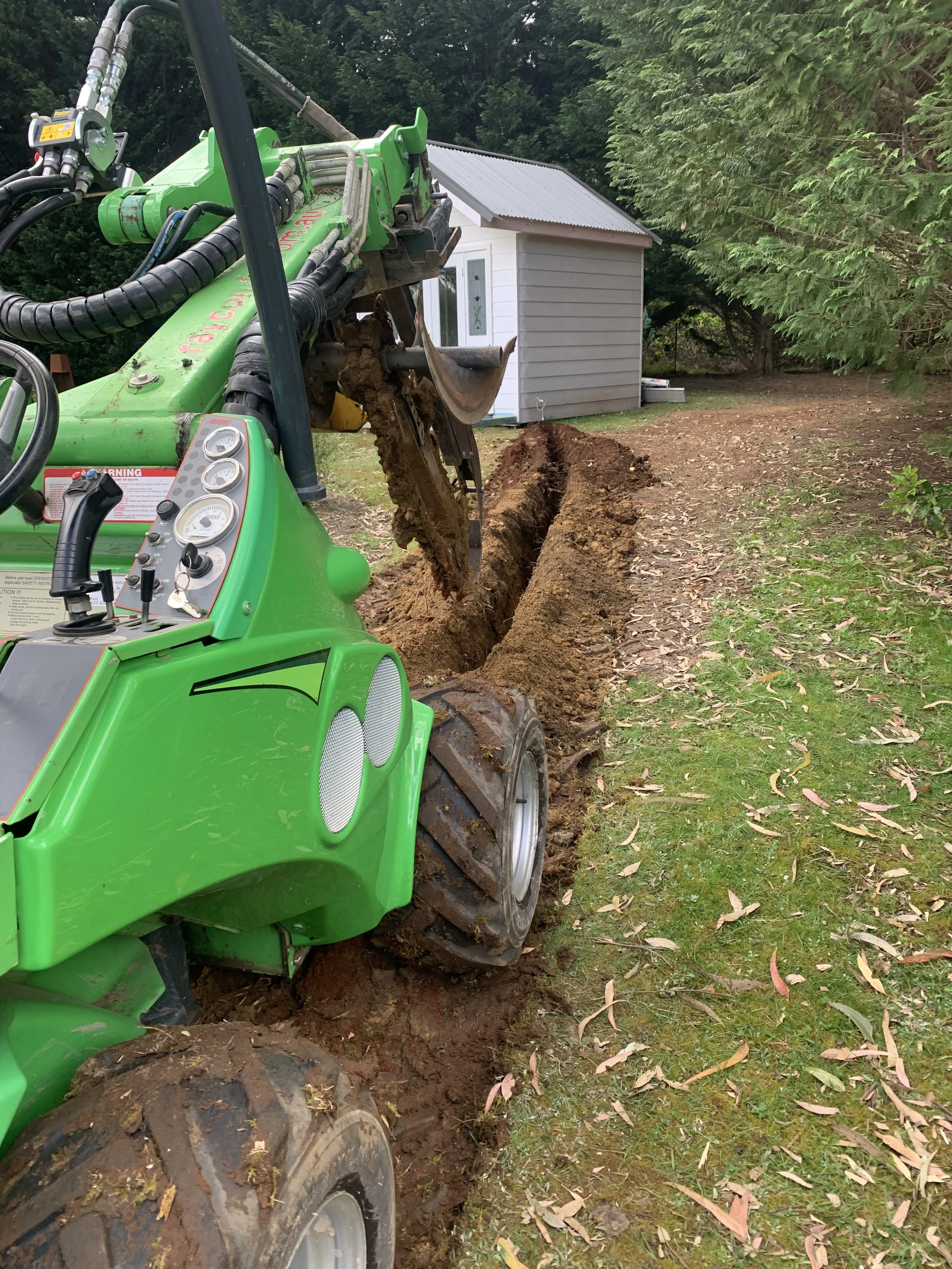 green Avant loader with trencher attachment digging a trench with white cottage in the background