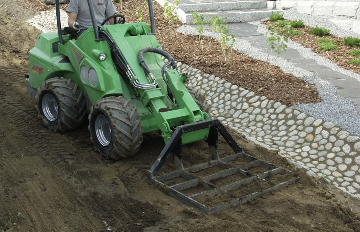 green Avant loader with the level bar attachment levlling soil with rock and walkway in the background