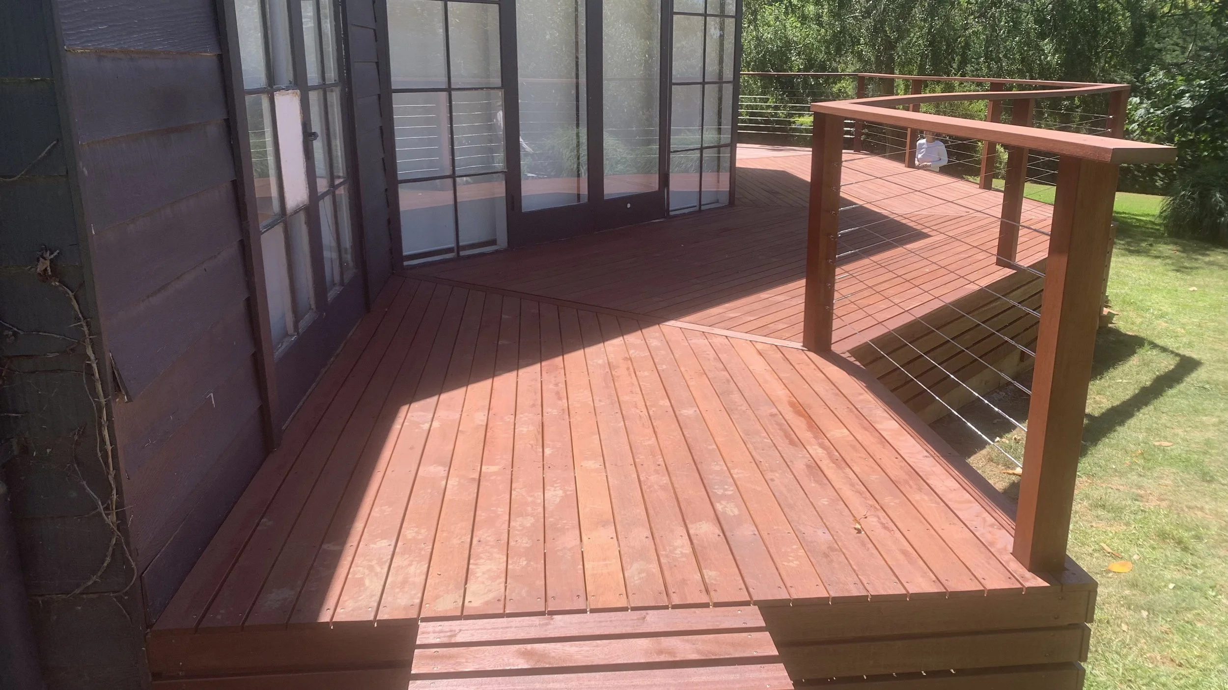 A wooden deck attached to a building with large glass doors and windows, surrounded by green trees and grass, with a person sitting on the lawn in the background.