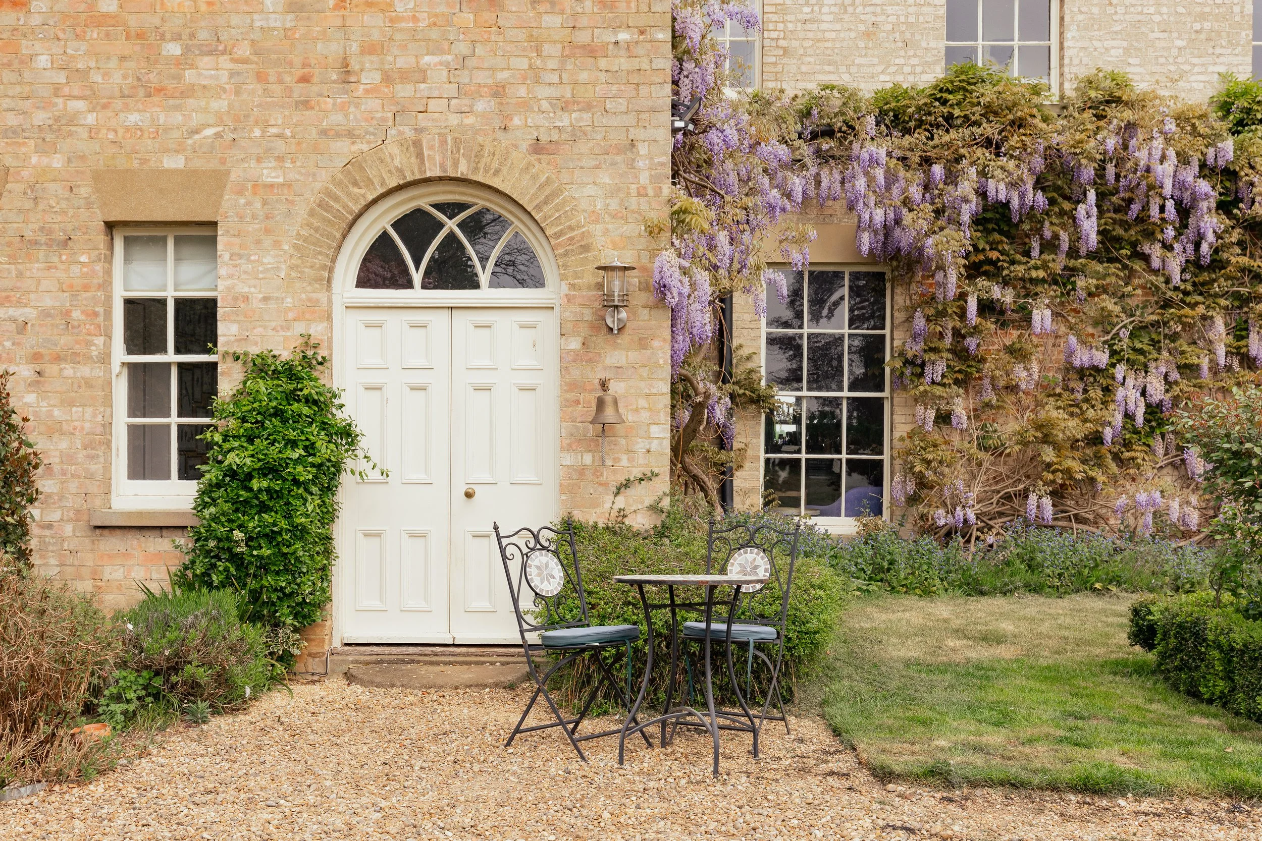 A cozy garden scene with a white door and two windows on a brick house. There is outdoor furniture with a small round table and two chairs on a gravel path. Purple flowers and ivy grow on the house walls, creating a lush, inviting atmosphere.