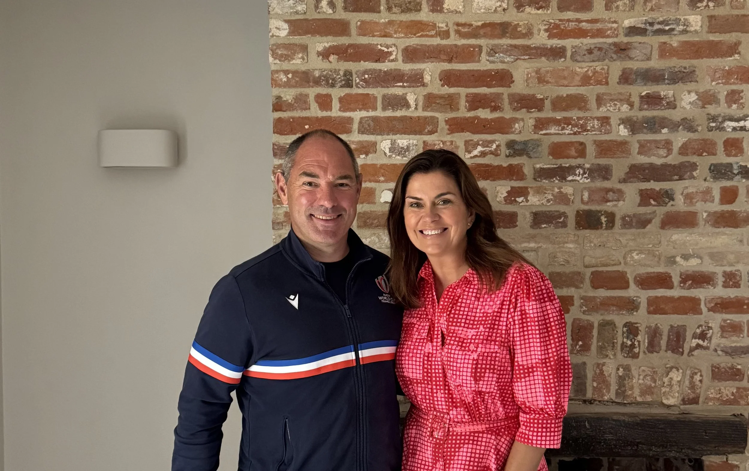 Phil Royle & TV star Amanda Lamb standing side by side in front of a brick wall, smiling at the camera. The man is wearing a navy blue jacket with red, white, and blue stripes, and the woman is wearing a red and pink checkered dress.