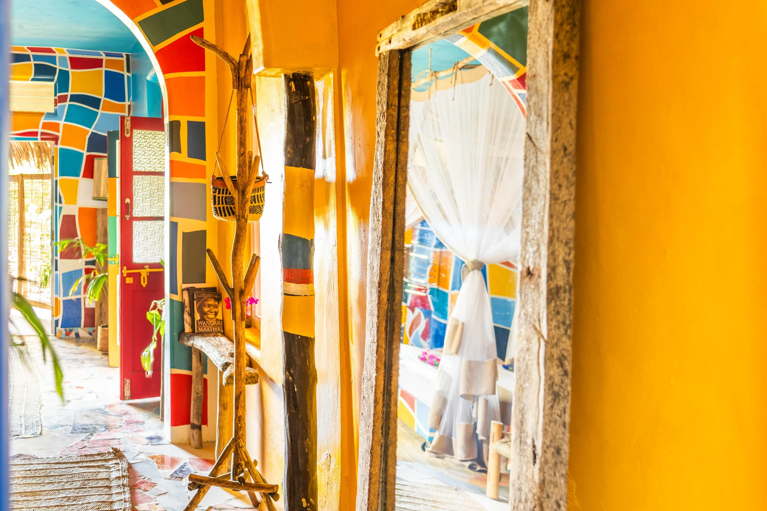 Colorful, eclectic interior with yellow walls, a colorful mosaic ceiling, a wooden coat rack, a door with a red frame, a mirror, and a window with white curtains in a home or cafe.
