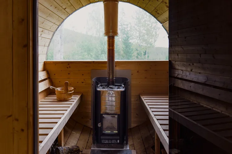 Inside a wooden sauna with two bench seats, a heater in the center with a chimney, and a wooden bucket and ladle on the left bench. Large window showing greenery outside.