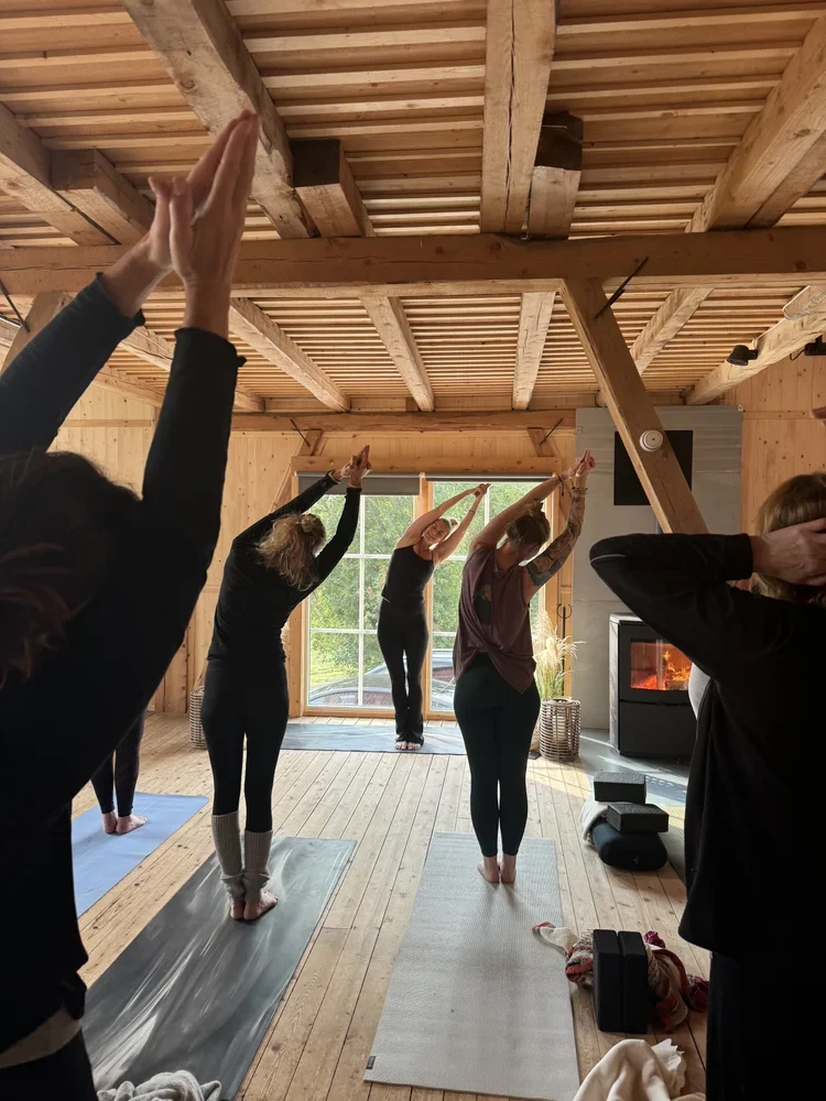 Group of people participating in a yoga class inside a wooden cabin, practicing a seated stretching pose with arms extended overhead.