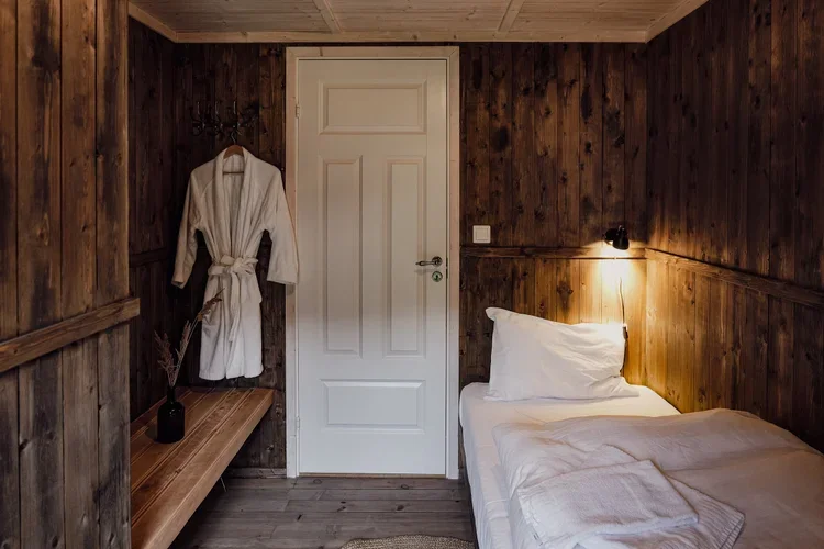 A rustic bedroom with wood-paneled walls, a white door, a single bed with white linens, a bathrobe hanging on a hook, and a small lamp on the wall.