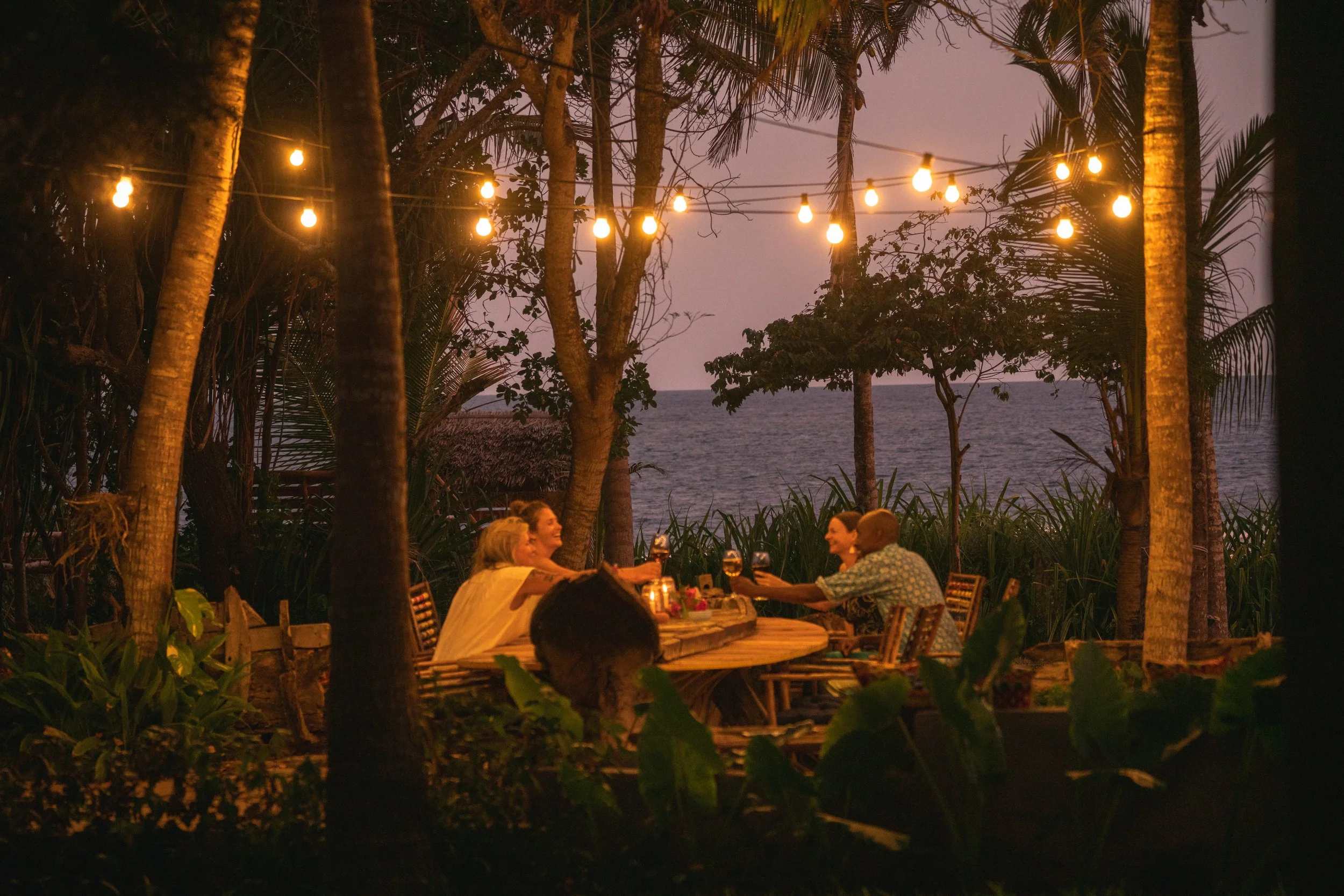 People enjoying a dinner outdoors at a tropical restaurant with string lights, palm trees, and ocean in the background at dusk.