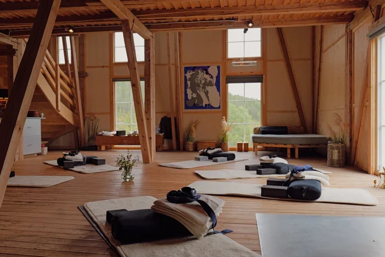 Yoga mats and folded blankets arranged on a wooden floor in a spacious, rustic studio with large windows, wooden beams, and minimal decor.
