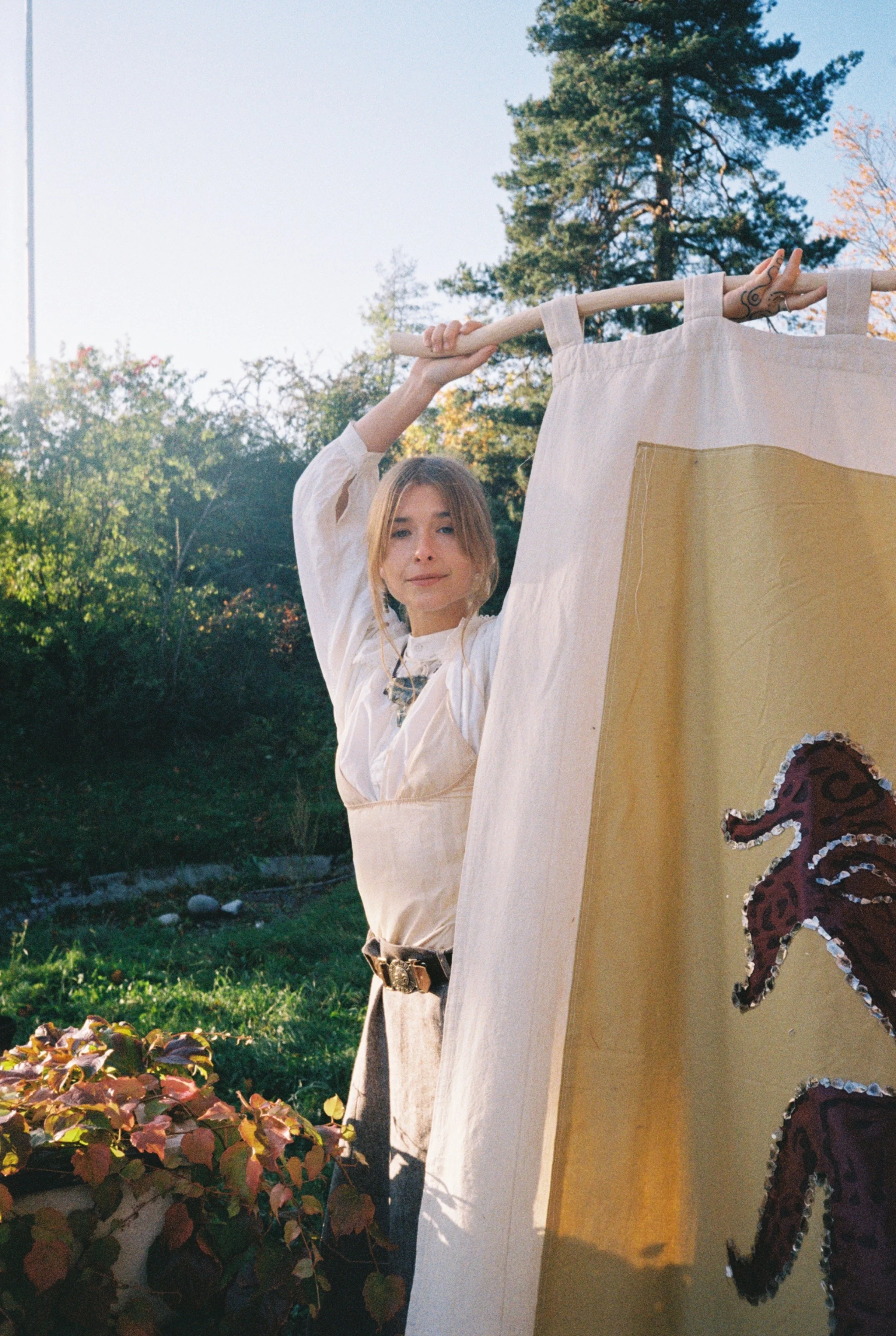 A woman in medieval costume standing outdoors next to a tent with a lion design, holding a pole. The background features trees and clear sky.