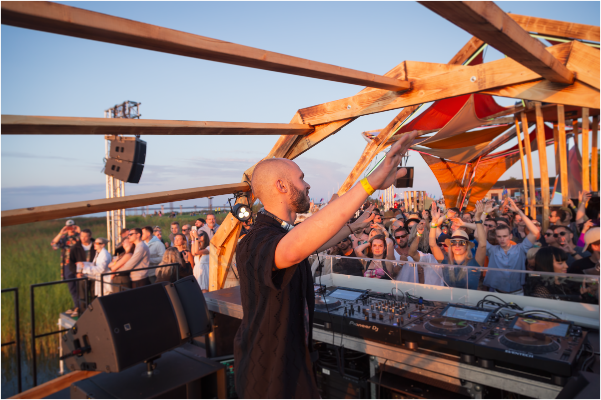 D J performing at an outdoor music festival during sunset with a crowd dancing in front of him. The structure is made of wooden beams and orange fabric, with speakers and DJ equipment visible.