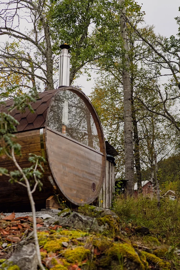 A small, round, wooden sauna with a glass window, surrounded by trees and moss-covered rocks in a natural outdoor setting.