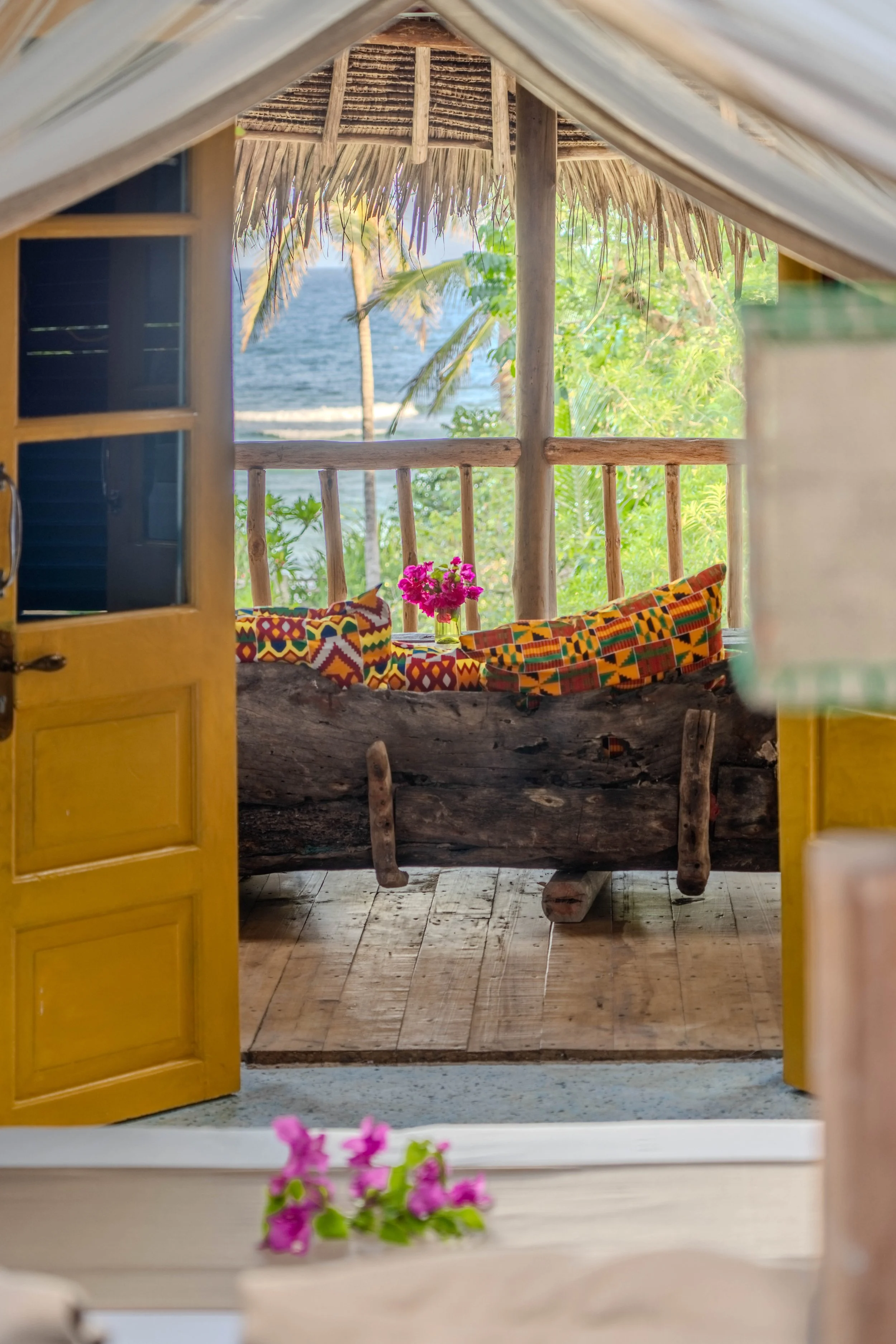 View through an open door showing a rustic porch with a wooden bench covered in colorful cushions, a vase with pink flowers, and a tropical landscape with palm trees and the ocean in the background.