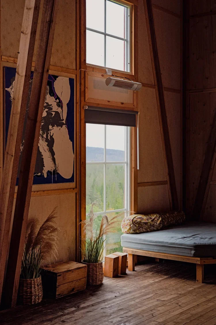 Interior of a rustic wooden cabin with two large windows showing a green landscape outside. There is a bed with a rolled blanket, potted plants, and a piece of art on the wall.