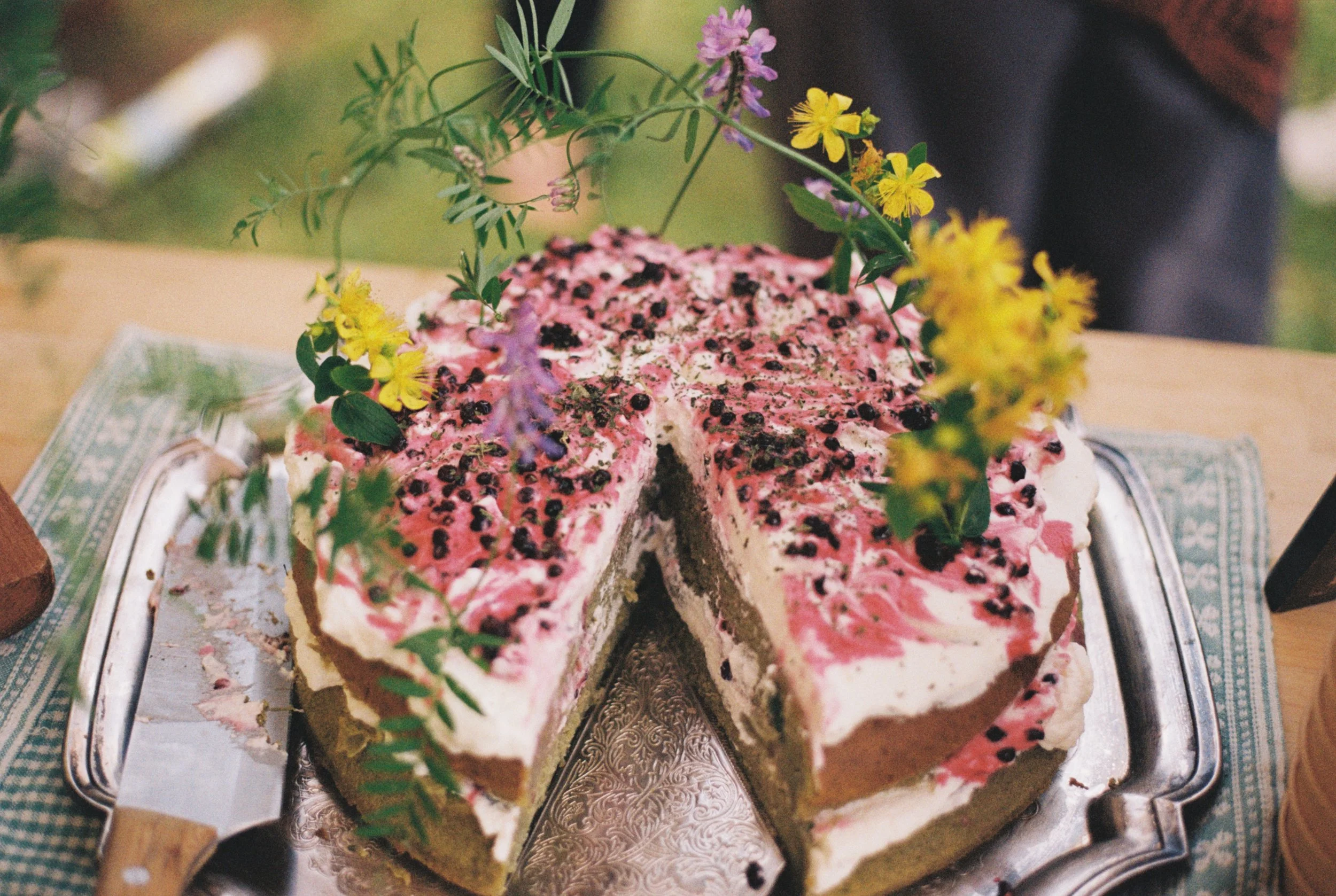 A partially sliced decorated cake with pink and white frosting, topped with small black berries and colorful flowers, placed on an ornate silver tray.