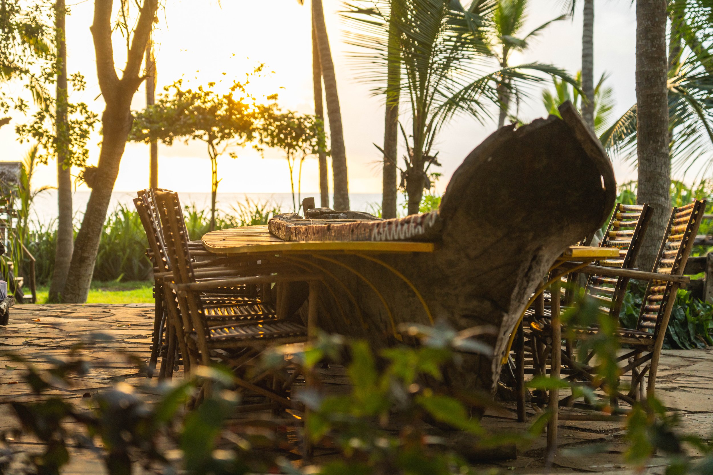 Outdoor dining area with a rustic wooden table and bamboo chairs, set amidst palm trees and tropical vegetation, overlooking a beach at sunset.
