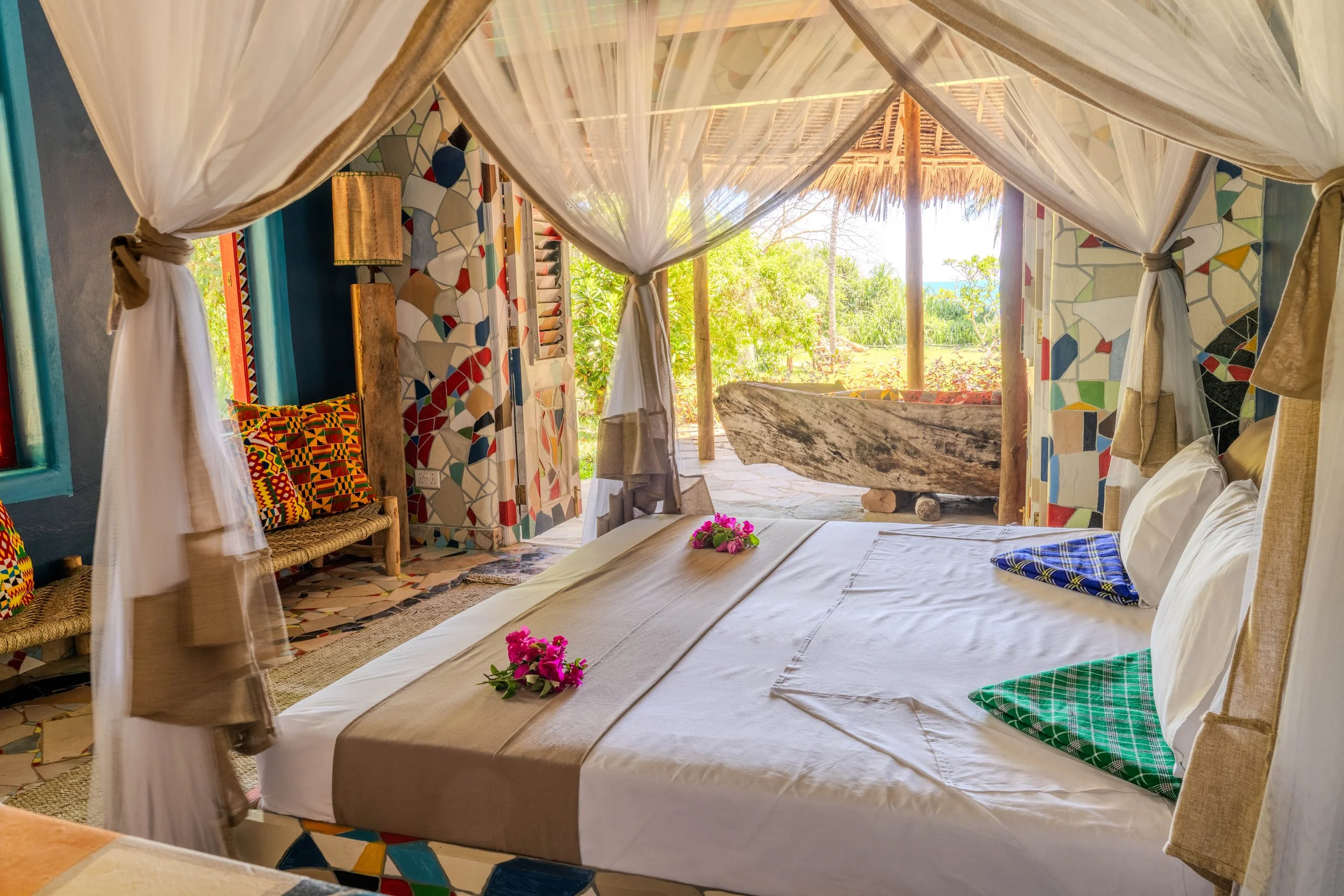 A cozy bedroom with a four-poster bed decorated with pink flowers, green and blue patterned pillows, and beige linens. The room features colorful mosaic walls, a window with teal framing, a wooden bench with patterned cushions, and an open view to a 