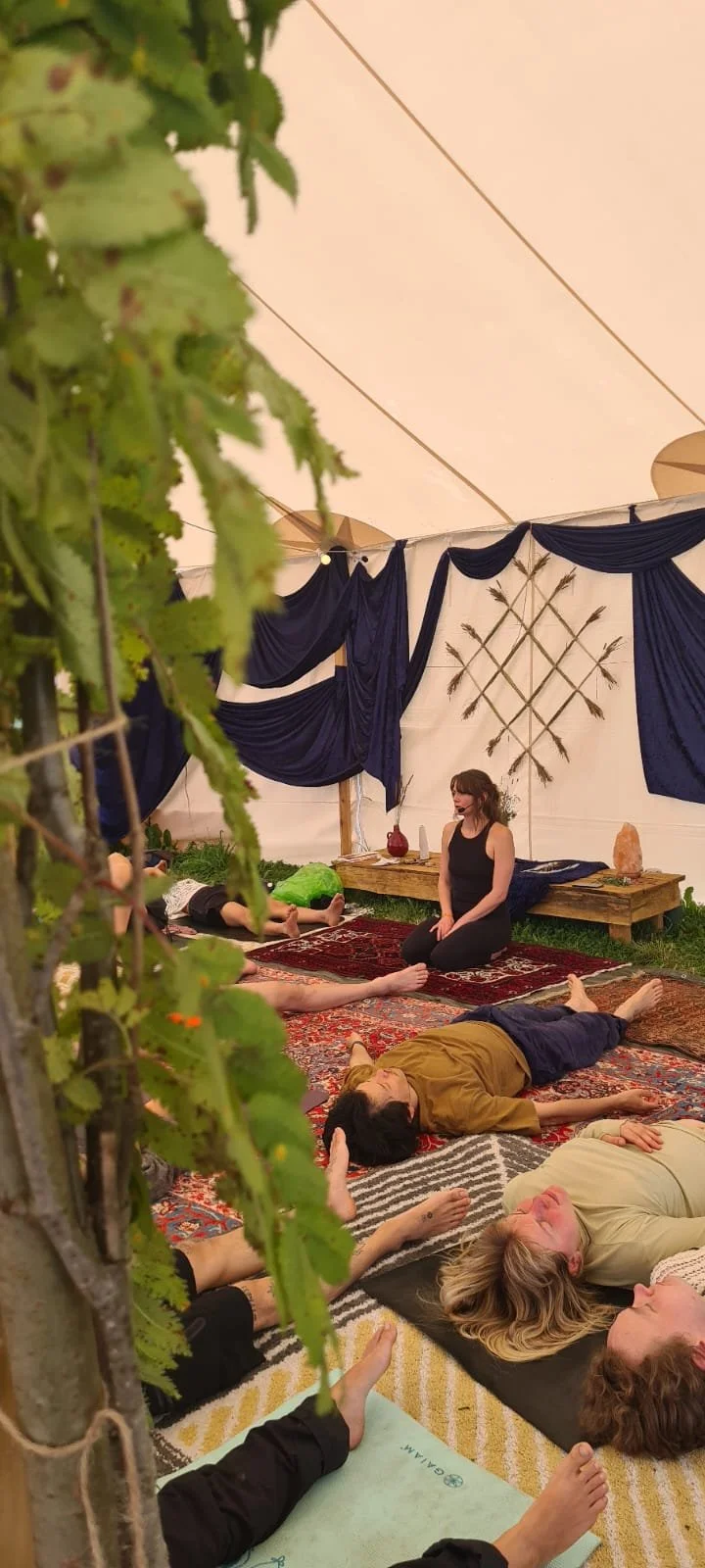 People participating in a yoga or meditation session inside a decorated tent with leafy green plants, rugs, and blue drapes on the walls.