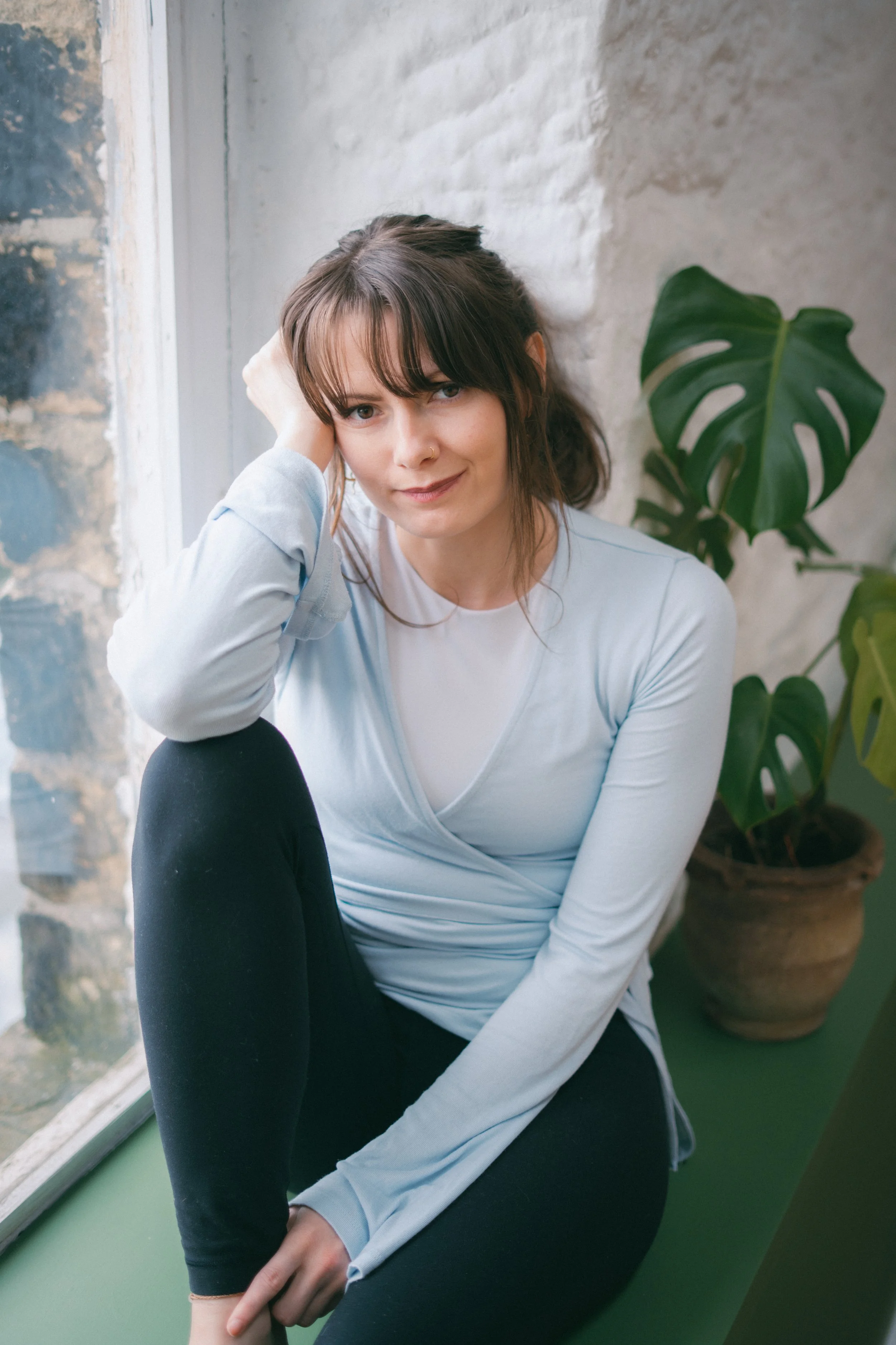 A woman with brown hair, wearing a light gray top, sitting by a window with a green plant in the background.