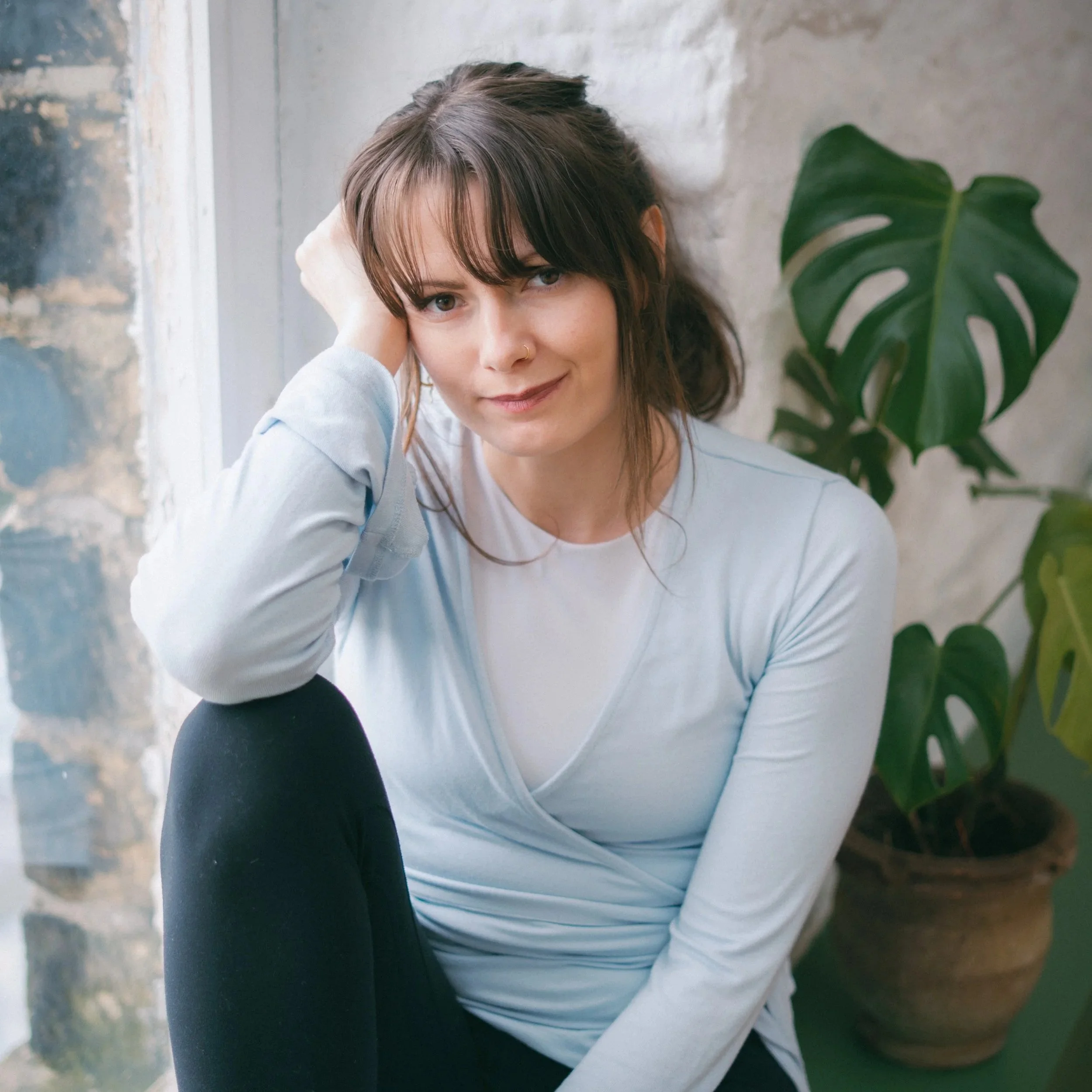 A woman with brown hair and bangs, wearing a light blue top, sitting indoors next to a large green monstera plant, with a stone wall and window in the background.