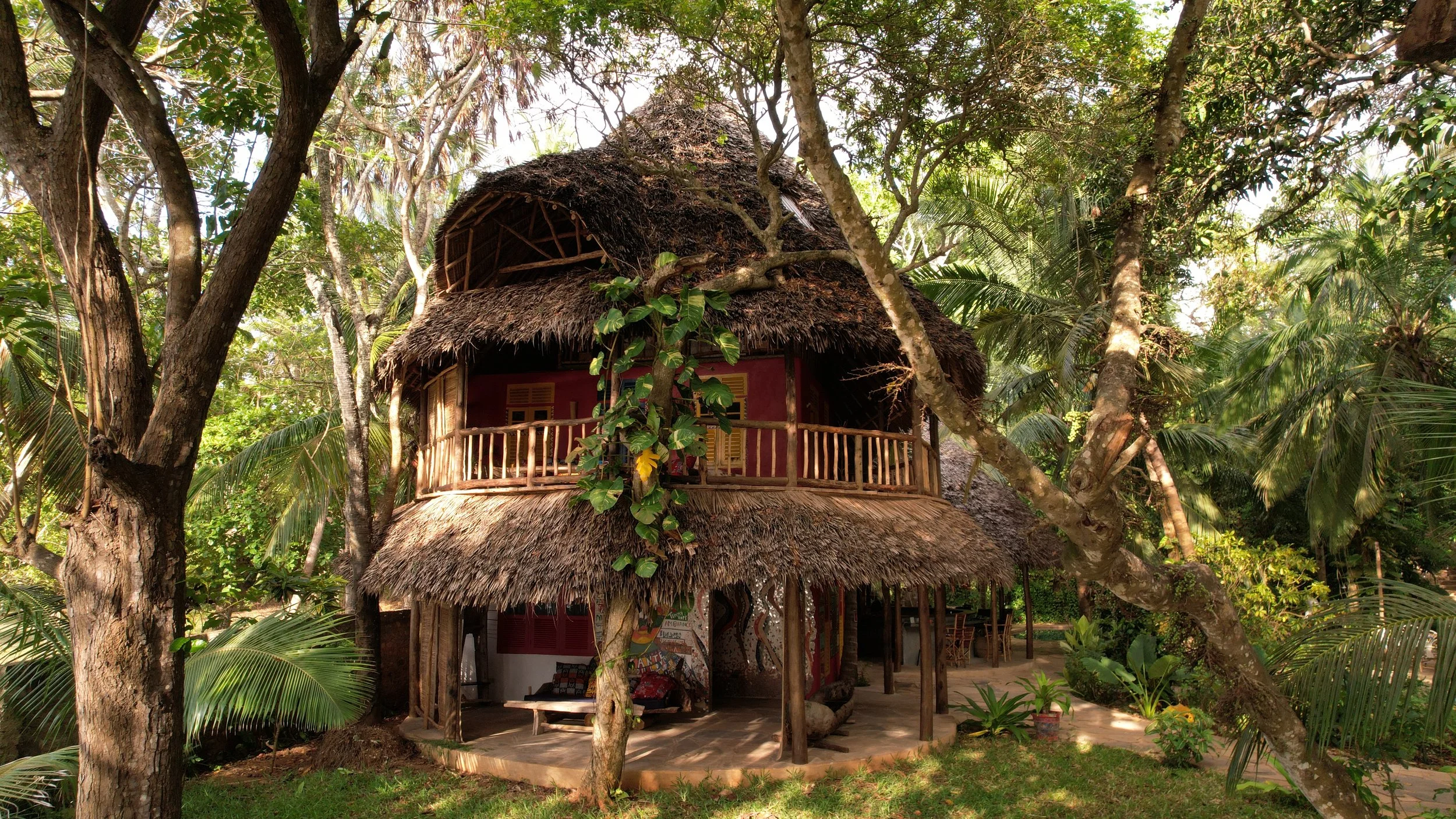 A two-story hut with a thatched roof surrounded by tropical trees and plants.