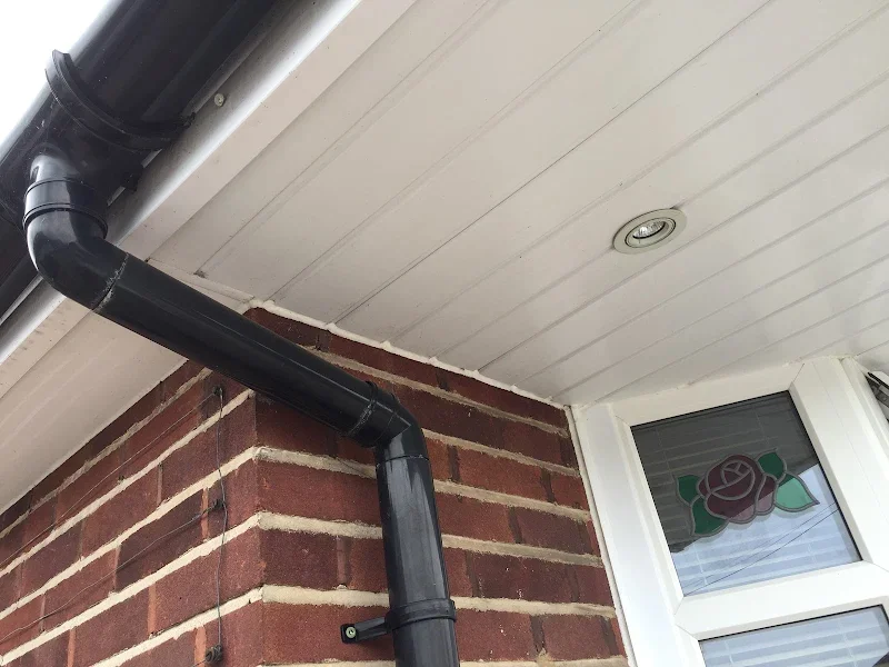 Close-up of a brick house corner with a black rain gutter pipe running down the side to the ground, under a white ceiling with a recessed light and a window with a stained glass rose flower design.