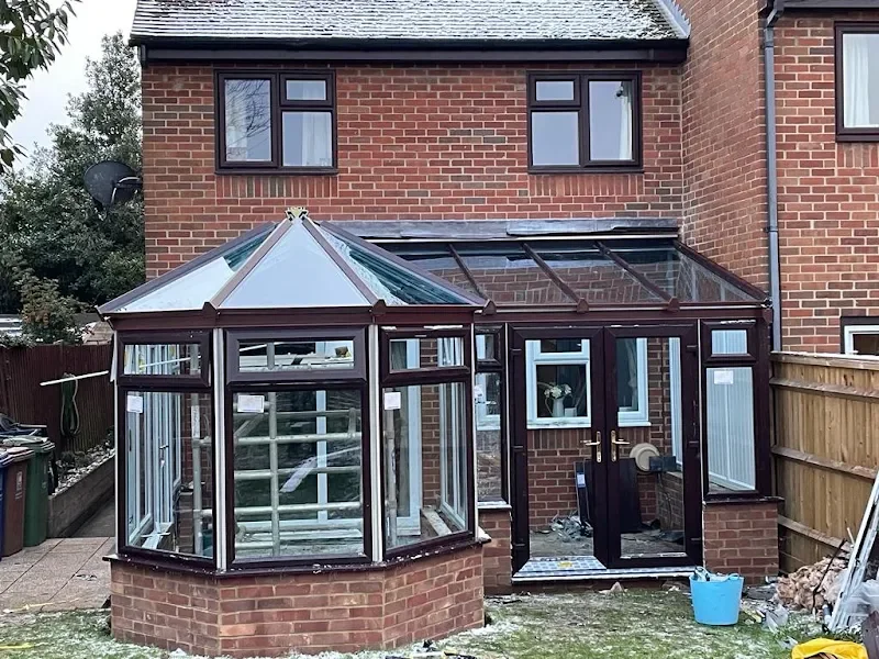 The image shows the back of a brick house with a wooden fence on one side, and an extension featuring a bay window and a porch with glass roofing. The extension has a glass door, and a satellite dish is mounted on the side of the house.