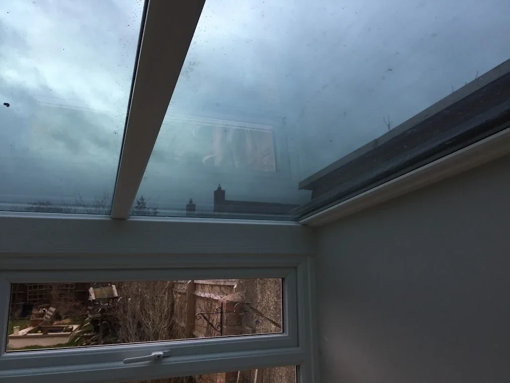 View through a large window with a glass ceiling, showing a cloudy sky and rooftops of neighboring houses.